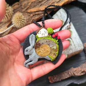 May include: A whimsical pendant necklace featuring a gray rabbit, a small house, and a fossilized ammonite. The pendant is accented with green moss, red berries, and a small green tree. The necklace has a black cord.