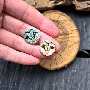 May include: Two small, round owl-themed ceramic pieces. One is light blue with black and orange details, the other is beige with black and orange details. The owl faces are centered on the pieces. The pieces are on a wooden surface.
