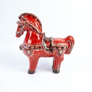 Op de afbeelding: Een rood keramiek paardenbeeldje met ingewikkelde details en een zadel. Het paard heeft een zwart hoofdstel en een bruin zadel met witte accenten.