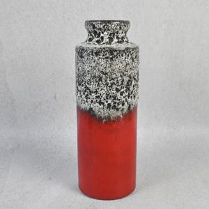 May include: A tall cylindrical ceramic vase featuring a vibrant red base that transitions into a textured, mottled black and white glaze at the top. The vase has a narrow neck and a flared rim, showcasing a distinct contrast between the smooth red finish and the rugged, speckled upper section of the vessel.