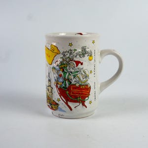 May include: White ceramic mug with a festive Christmas design. The mug features a colourful illustration of Santa Claus in a sleigh, with stars and text accents. The mug has a curved handle.