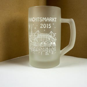 May include: Frosted glass beer stein with a handle. The stein features a white graphic of a Christmas market scene and the text "NACHTSMARKT 2015". The glass is cylindrical with a slightly flared base.