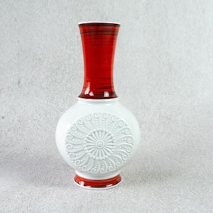 Könnte beinhalten: Weiße Keramikvase mit einem rot-schwarz gestreiften Hals und einem weißen, geprägten Blumendesign. Die Vase ist rund mit einem breiten Boden und einem schmalen Hals.