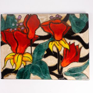 Könnte beinhalten: Eine Keramikfliese mit einem floralen Design. Die Fliese zeigt drei rote Blumen mit gelben Zentren und grünen Blättern auf einem weißen Hintergrund mit schwarzen Linien.