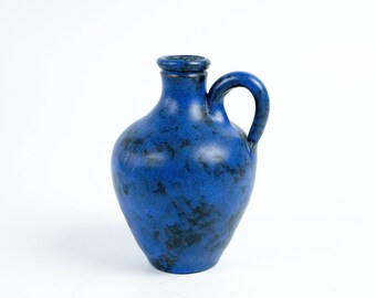 May include: A blue ceramic pitcher with a handle. The pitcher has a textured surface with dark blue and black speckles. The handle is arched, and the opening is small. The pitcher is a decorative item.