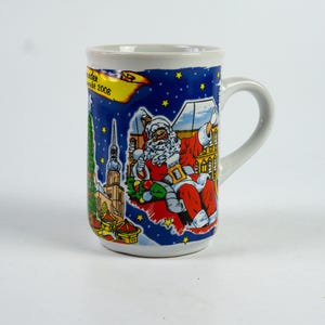 May include: White ceramic mug with a blue background featuring Santa Claus, buildings, and stars. The mug has a curved handle and a colourful design, with text that reads "Wunder Markt 2008".