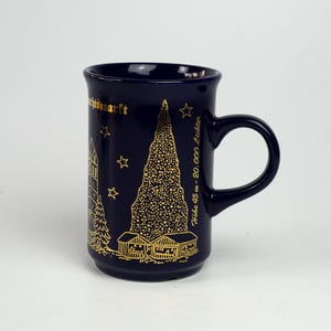May include: A dark blue ceramic mug with a gold design featuring a Christmas tree, buildings, stars, and text. The mug has a handle and is likely for hot beverages. The design includes the text "Höhe 45 m - 20.000 Lichter".
