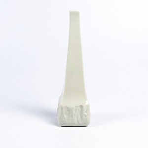 May include: A white ceramic vase with a tall, narrow, triangular shape and a textured base.