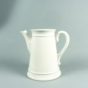 May include: A white ceramic pitcher with a handle and a spout. The pitcher has a simple design with a rounded body and a flared base.