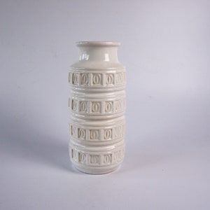May include: White ceramic vase with a geometric pattern. The vase has a cylindrical shape and is decorated with raised circles and squares.