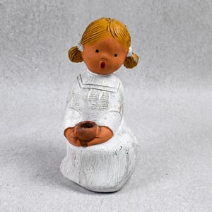 May include: A ceramic figurine of a girl with blonde pigtails, wearing a white dress, and holding a brown cup. The figurine has a surprised expression and is painted with a distressed finish. The overall theme is a decorative item.