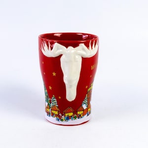 May include: Red ceramic mug with a white moose head design. The mug features a winter village scene around the base and gold stars. The word "Welcome" is visible on the side of the mug.