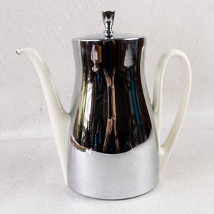 May include: A vintage coffee pot with a shiny chrome finish and a white handle and spout. The pot has a lid with a small decorative handle. The design is a mix of chrome and white ceramic.