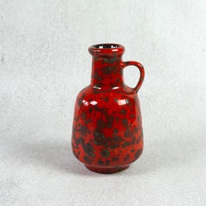 May include: A red ceramic vase with a speckled glaze and a handle. The vase is about 10 inches tall and has a round body.