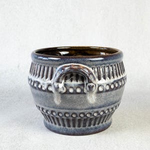 May include: A gray ceramic planter with a dark brown interior and a handle. The planter features a textured design with vertical lines and dotted patterns. The planter is suitable for small plants or decorative use.