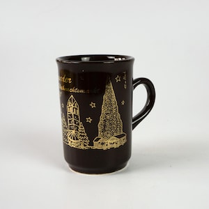 May include: A dark brown ceramic mug featuring a gold illustration of a building, trees, and text. The mug has a curved handle and a cylindrical shape. The text on the mug appears to be in German.