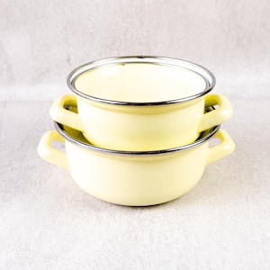 Può includere: Due ciotole in smalto giallo chiaro impilate. Ogni ciotola ha un bordo argentato e due piccole maniglie. Le ciotole sono di colore giallo tenue. Ideali per cucinare o servire.