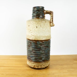 May include: A tall, ceramic vase with a blue and brown glaze. The vase has a wide mouth and a single handle. The vase is decorated with a textured pattern.