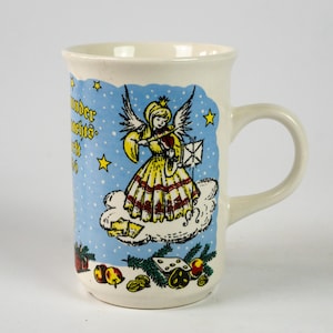 May include: Cream-colored ceramic mug with a handle, featuring a whimsical illustration. The design includes a winged angel playing a violin on a cloud, surrounded by stars and text. Festive elements like fruit and nuts adorn the base.