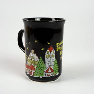 May include: Black ceramic mug with a handle, featuring a colourful illustration of a European village scene with buildings, trees, and a carousel. The mug has gold text and yellow star accents. A festive holiday-themed design.
