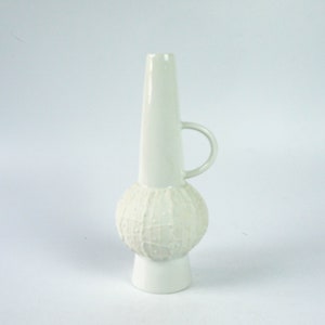May include: White ceramic vase with a textured bulbous base and a tall, slender neck. The vase has a single handle.