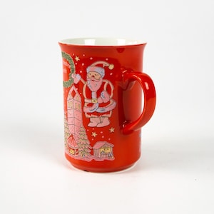 May include: A red ceramic mug with a white interior and a large red handle. The mug features a colourful illustration of Santa Claus, a church, and a Christmas wreath. The mug is decorated with stars and a small house.