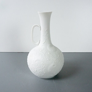 May include: White porcelain vase with a floral design. The vase has a round body and a long, thin neck with a handle.