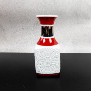 May include: A decorative vase with a white, red, and silver design. The vase has a square base and a flared top. The white section features an embossed floral pattern. The top and base are red, and the neck is silver.