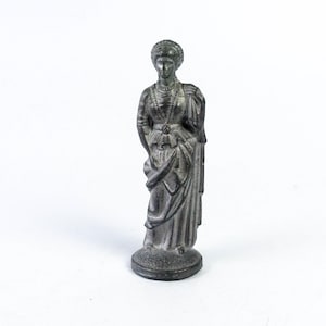Può includere: Una piccola statuetta di metallo grigio di una donna con un lungo vestito e una corona. La statuetta è in piedi su una piccola base.