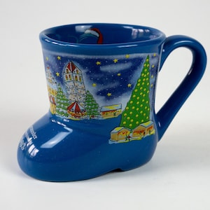 May include: A blue ceramic boot-shaped mug with a handle. The mug features a winter scene with a Christmas tree, buildings, and stars. The mug has a glossy finish and is designed for hot beverages.