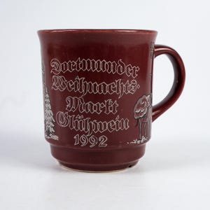 May include: A burgundy ceramic mug with a handle, featuring white lettering and illustrations. The text on the mug reads "Dortmunder Weihnachts Markt Glühwein 1992". The mug has a capacity of 0.2 liters.