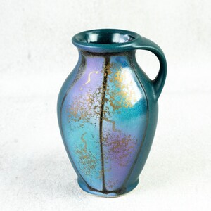 May include: A teal ceramic vase with a handle, displaying a gradient of blue and purple tones. The decorative vase has gold accents and a unique shape. Ideal for displaying flowers or as a standalone art piece.
