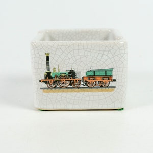 以下が含まれることがあります： ひび割れた釉薬仕上げの白いセラミック製の小物入れ。箱には、蒸気機関車が貨車を引くヴィンテージのイラストが描かれています。
