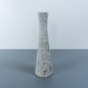 May include: A white ceramic vase with a textured surface. The vase has a tall, slender shape and a narrow opening.