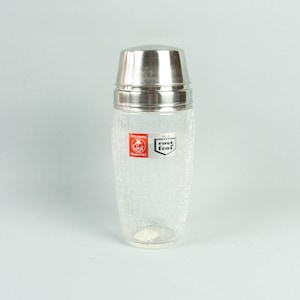 May include: A vintage cocktail shaker with a clear, textured glass body and a shiny, silver-colored metal lid. The shaker has a crackle pattern and a label with red and white text. The overall design is simple and classic.