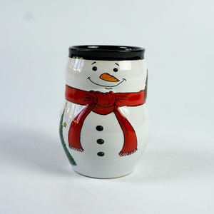 Könnte beinhalten: Weißer Keramikbecher in Schneemannform mit schwarzem Rand und rotem Schal. Der Schneemann hat eine Karottennase, schwarze Augen und einen lächelnden Mund. Auf der Seite ist ein kleiner Weihnachtsbaum aufgemalt.