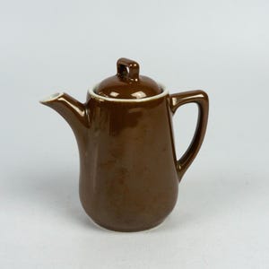 May include: A brown ceramic creamer with a white rim and a white lid. The creamer has a rounded body and a handle.