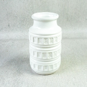 May include: A white ceramic vase with a cylindrical body and a narrow neck. The vase has a repeating geometric pattern around its body. The vase is a solid white colour and has a glossy finish. The vase is suitable for displaying flowers or as a decorative item.