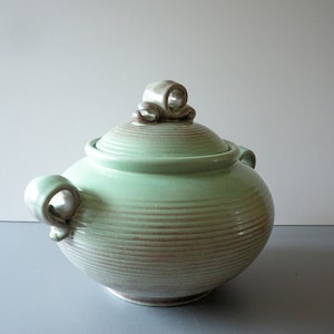 Può includere: Un piatto da portata in ceramica verde con coperchio. Il coperchio ha una maniglia decorativa. Il piatto ha un motivo a strisce marroni e verdi.