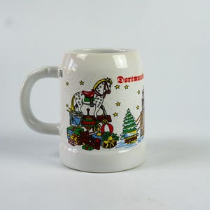 May include: White ceramic beer stein with a handle, featuring a colorful illustration of a rocking horse, wrapped gifts, a Christmas tree, and the word "Dortmund". The stein has a wide base and a slightly tapered top.