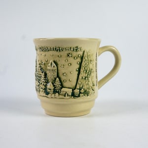 May include: A beige ceramic mug with a handle, featuring a raised relief design of a winter village scene with a church, trees, and the text "Weihnachtsmarkt". The design is in shades of green and the mug has a slightly flared shape.