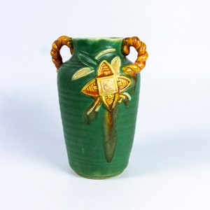 Könnte beinhalten: Eine grüne Keramikvase mit einem geometrischen Muster in Gelb- und Orangetönen. Die Vase hat zwei gedrehte Seilgriffe.