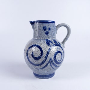 Può includere: Un'anfora in ceramica blu e bianca con una superficie testurizzata e un design a spirale. L'anfora ha un manico e un beccuccio.