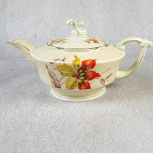 Könnte beinhalten: Cremefarbene Keramik-Teekanne mit einem Blumendesign in Gelb, Orange und Braun. Die Teekanne hat einen gebogenen Ausguss, einen Schlaufengriff und einen Deckel mit dekorativem Knauf. Das Blumenmuster wiederholt sich auf dem Deckel.