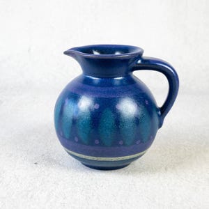 May include: A small, round, blue ceramic pitcher with a handle and spout. The pitcher features a dark blue glaze with teal and purple accents, including a band of dots and vertical patterns. The handle is curved and the spout is small.