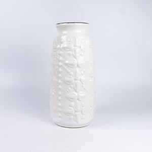 May include: White ceramic vase with an embossed floral design. The vase has a tall, cylindrical shape and a smooth, white finish.