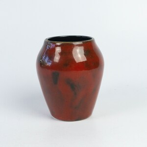 Può includere: Un piccolo vaso in ceramica rosso con un bordo nero. Il vaso ha una finitura lucida e un motivo screziato.