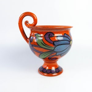 May include: A ceramic cup with a unique, curled handle and a footed base. The cup is decorated with a colorful floral design in shades of orange, blue, green, and purple.