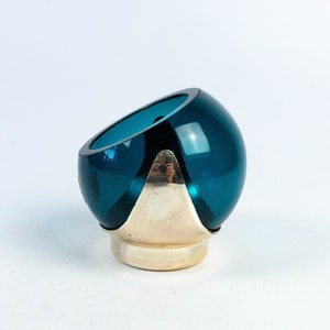 May include: A teal-colored glass bowl with a silver-colored base. The bowl has a rounded shape with a slanted opening. The base is a combination of a circular base and a triangular support.