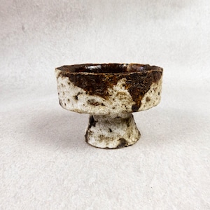 Può includere: Una piccola ciotola in ceramica fatta a mano con base a piedistallo. La ciotola presenta un esterno strutturato bianco sporco con accenti marroni e un interno marrone scuro. Il design è rustico e organico, adatto all'uso decorativo.